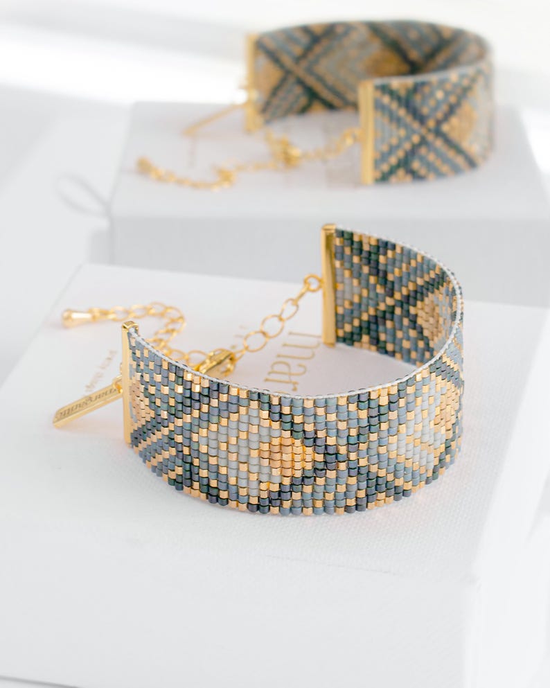 Op de afbeelding: Een kralenarmband met een geometrisch patroon in tinten grijs, goud en blauwgroen. De armband heeft goudkleurige metalen accenten en een kettingsluiting. Een andere armband is zichtbaar op de achtergrond.