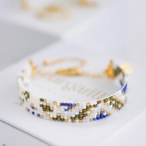 May include: A beaded bracelet with a white base, featuring a pattern of blue, green, and gold beads. The bracelet is displayed on a white surface, with a blurred bracelet in the background. The bracelet has a gold clasp.