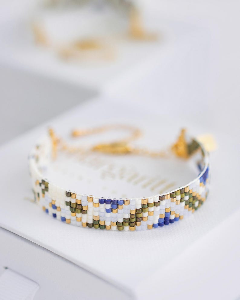 May include: A beaded bracelet with a white base, featuring a pattern of blue, green, and gold beads. The bracelet is displayed on a white surface, with a blurred bracelet in the background. The bracelet has a gold clasp.