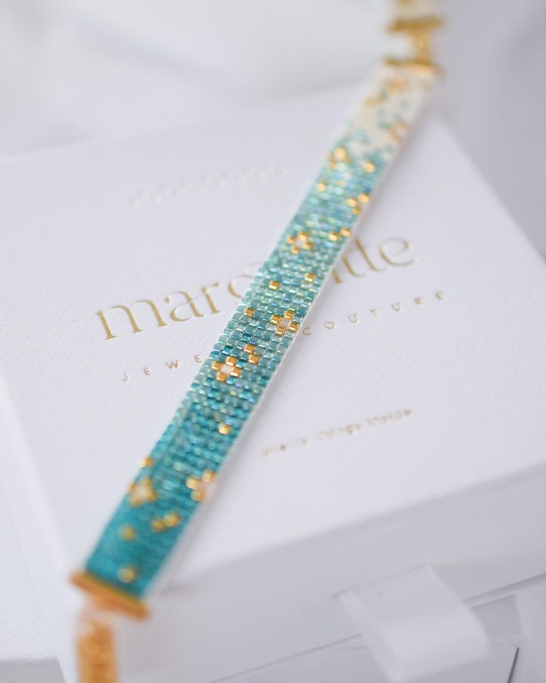 May include: A beaded bracelet with a gradient of turquoise and gold beads. The bracelet is on a white box with the words "marcade JEWELRY COUTURE" embossed in gold. The bracelet's design features a pattern of small, square beads.