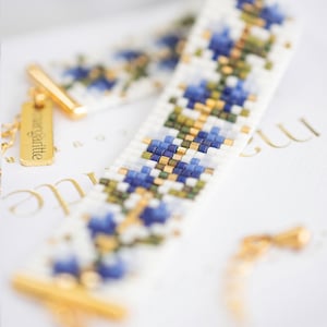 May include: A beaded bracelet with a floral pattern in shades of blue, white, green, and gold. The bracelet has a gold clasp and a small gold tag that reads "margritte". The bracelet is lying on a white surface.