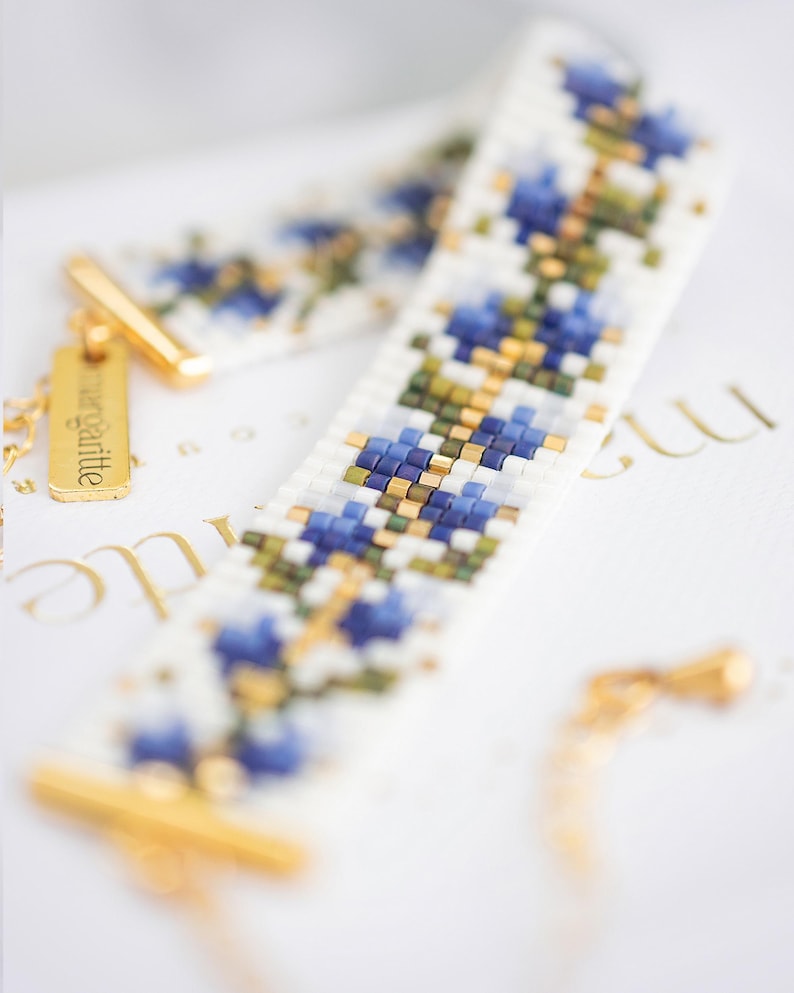 May include: A beaded bracelet with a floral pattern in shades of blue, white, green, and gold. The bracelet has a gold clasp and a small gold tag that reads "margritte". The bracelet is lying on a white surface.