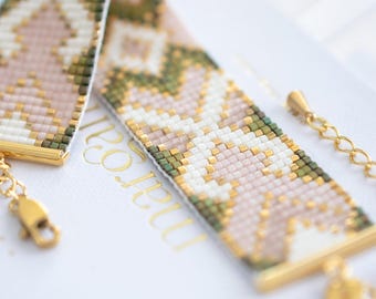 May include: A beaded bracelet with a geometric pattern in shades of pink, white, green, and gold. The bracelet has gold-colored metal accents and a chain closure. The bracelet is resting on a white surface.