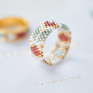 May include: A beaded ring with a geometric pattern in white, gold, pink, and teal. The ring is in focus, with a blurred gold ring in the background. The ring's design features small beads arranged in a repeating pattern.