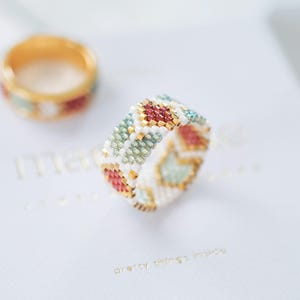 May include: A beaded ring with a geometric pattern in red, gold, white, and teal. The ring is in focus, with a second ring in the background. The rings are on a white surface with gold text that reads "pretty things inside".