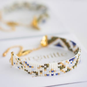 May include: A beaded bracelet with a geometric pattern in white, blue, gold, and olive green. The bracelet is on a white surface, with a second bracelet slightly out of focus in the background. The bracelet has a gold clasp.