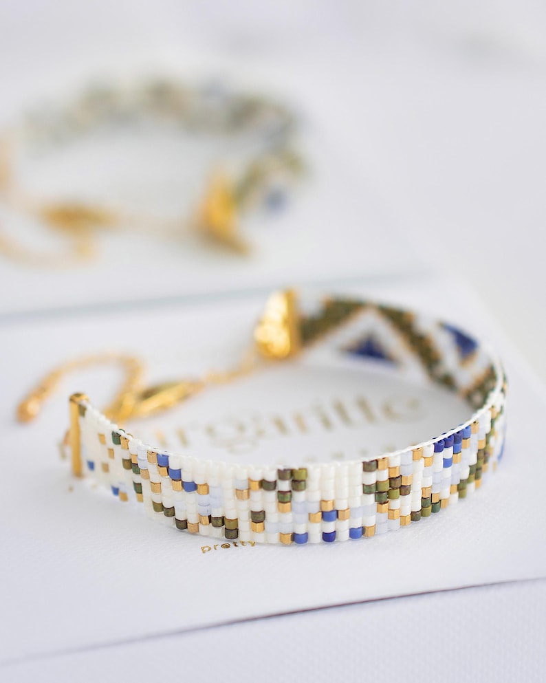 May include: A beaded bracelet with a geometric pattern in white, blue, gold, and olive green. The bracelet is on a white surface, with a second bracelet slightly out of focus in the background. The bracelet has a gold clasp.