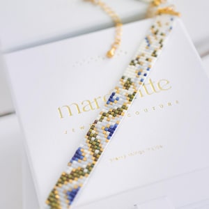 May include: A beaded choker necklace with a white base, featuring a pattern of blue, green, and gold beads. The necklace is displayed on a white box with gold text that reads "margette JEWELRY COUTURE Pretty things inside."