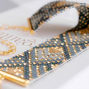 Op de afbeelding: Een kralenarmband met een geometrisch patroon in goud, teal en grijs. De armband heeft goudkleurige metalen eindkappen en een gouden ketting. Het ingewikkelde kralenwerk cre&euml;ert een textuur, wat het vakmanschap van de sieraden laat zien.