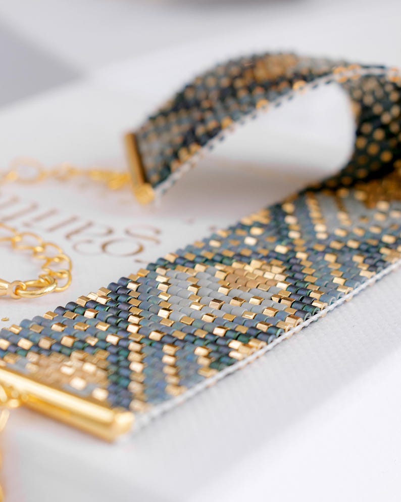 Op de afbeelding: Een kralenarmband met een geometrisch patroon in goud, teal en grijs. De armband heeft goudkleurige metalen eindkappen en een gouden ketting. Het ingewikkelde kralenwerk cre&euml;ert een textuur, wat het vakmanschap van de sieraden laat zien.