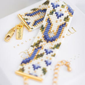 May include: A beaded bracelet with a white background, featuring a geometric pattern in blue, green, and gold. The bracelet has gold-colored metal accents and a small tag that reads "margalitte". The bracelet is a stylish accessory.