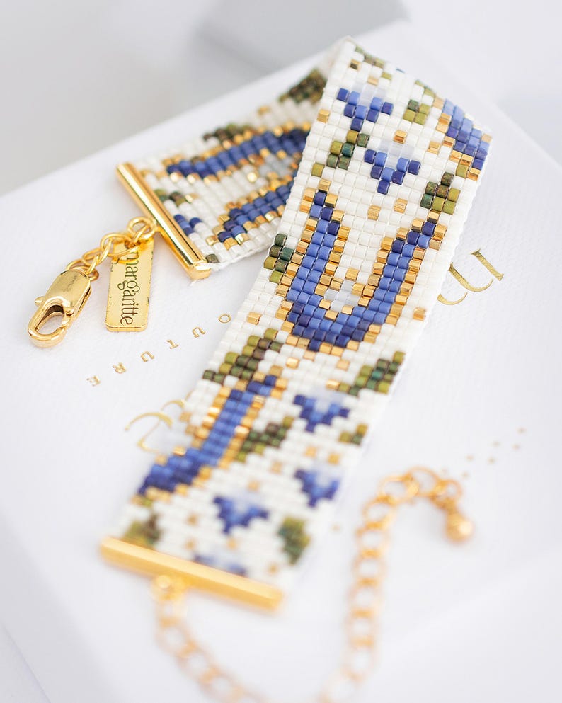 May include: A beaded bracelet with a white background, featuring a geometric pattern in blue, green, and gold. The bracelet has gold-colored metal accents and a small tag that reads "margalitte". The bracelet is a stylish accessory.