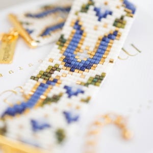 May include: Close-up of a beaded bracelet with a white background. The bracelet features a geometric pattern in blue, green, and gold beads. The clasp and small tag are gold. The bracelet is approximately 0.75 inches wide.