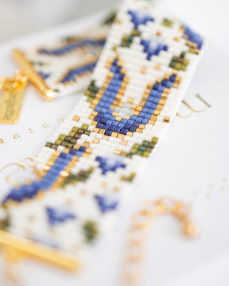 May include: Close-up of a beaded bracelet with a white background. The bracelet features a geometric pattern in blue, green, and gold beads. The clasp and small tag are gold. The bracelet is approximately 0.75 inches wide.