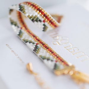 Puede incluir: Una pulsera de cuentas con un patrón geométrico en blanco, verde, rojo y dorado. La pulsera está sobre una superficie blanca con texto dorado. El cierre y la cadena son dorados.