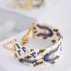 May include: A beaded bracelet with a white base, featuring a pattern of blue, green, and gold beads. The bracelet has a gold clasp and chain. The design is intricate and geometric, with a focus on color and texture. Another bracelet is slightly visible in the background.