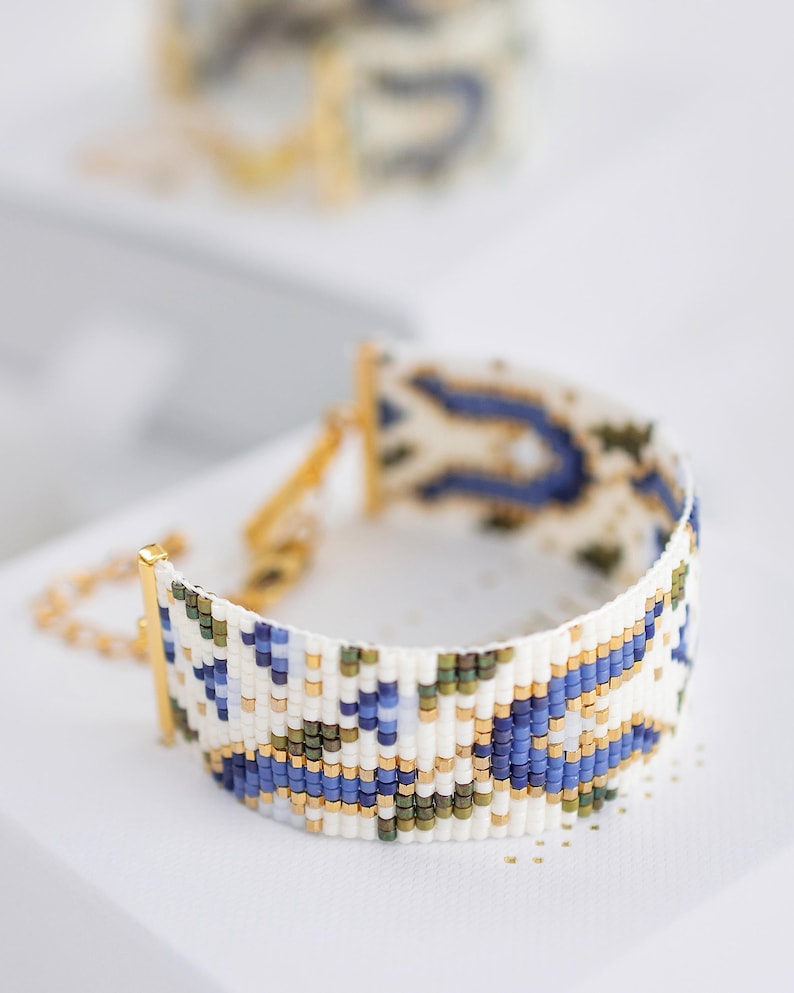 May include: A beaded bracelet with a white base, featuring a pattern of blue, green, and gold beads. The bracelet has a gold clasp and chain. The design is intricate and geometric, with a focus on color and texture. Another bracelet is slightly visible in the background.