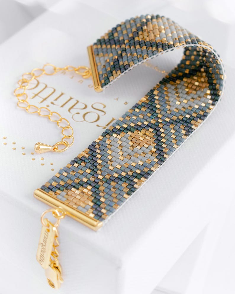 Op de afbeelding: Een kralenarmband met een geometrisch patroon in grijstinten, goud en donkergroen. De armband heeft goudkleurige metalen eindkappen en een bijpassende ketting sluiting. De merknaam "marguerite" is op de sluiting gegraveerd.