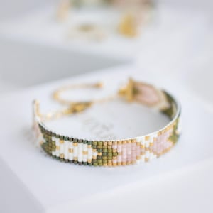 Op de afbeelding: Een kralenarmband met een gouden ketting en sluiting. De armband heeft een patroon van witte, roze, groene en gouden kralen. De armband ligt op een wit oppervlak.