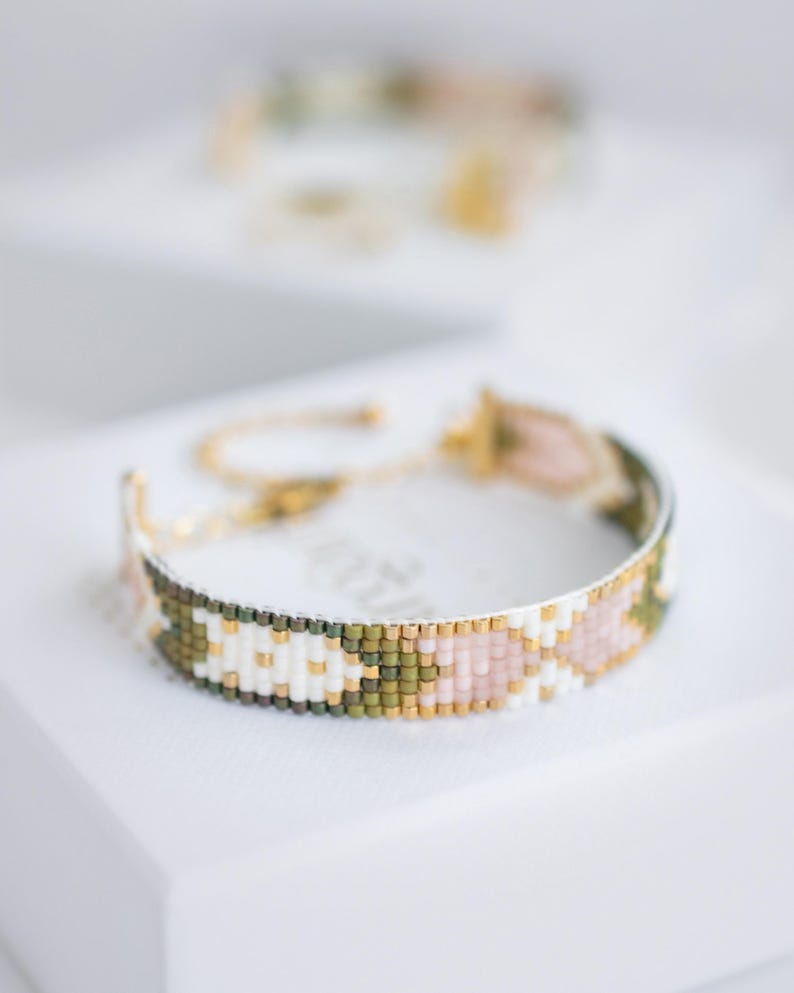Op de afbeelding: Een kralenarmband met een gouden ketting en sluiting. De armband heeft een patroon van witte, roze, groene en gouden kralen. De armband ligt op een wit oppervlak.