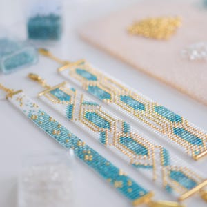 May include: Three beaded bracelets with gold clasps and accents. The bracelets feature white, turquoise, and gold seed beads in geometric patterns. The bracelets are displayed on a white surface with other crafting supplies in the background.