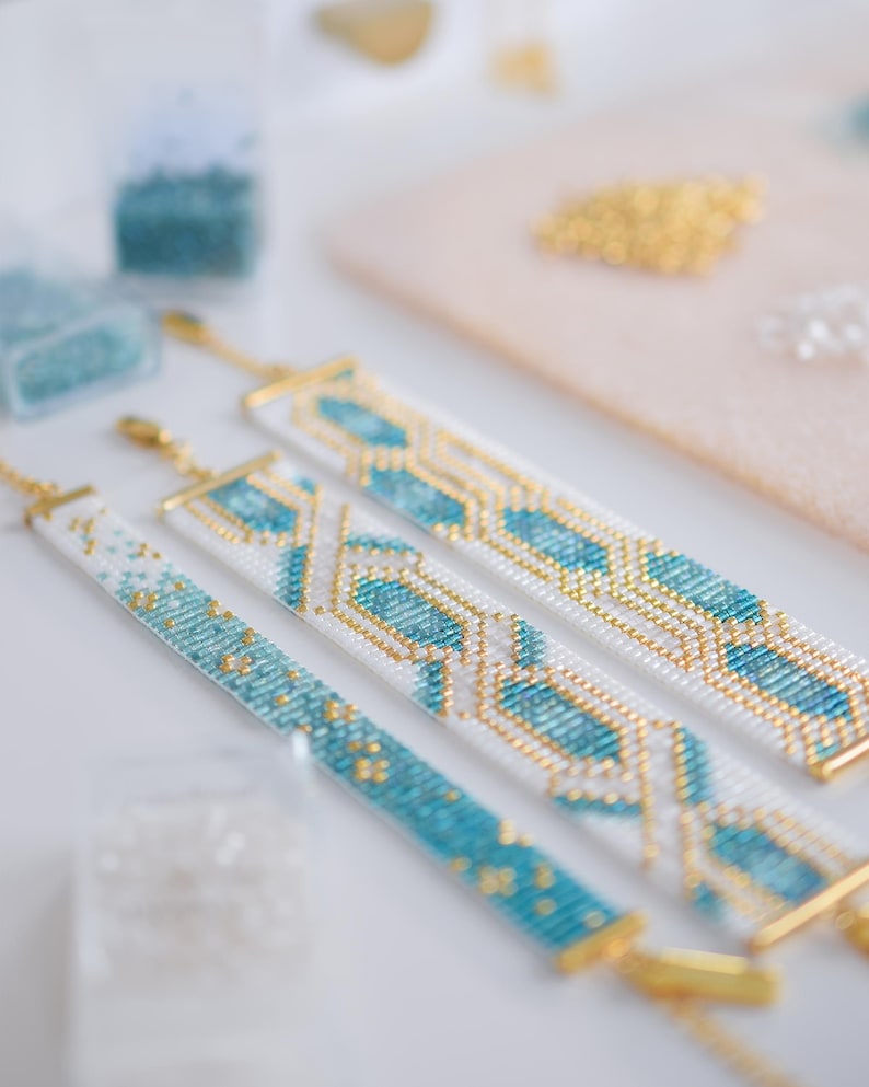 May include: Three beaded bracelets with gold clasps and accents. The bracelets feature white, turquoise, and gold seed beads in geometric patterns. The bracelets are displayed on a white surface with other crafting supplies in the background.