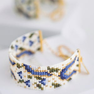 May include: A beaded bracelet with a white base, featuring a geometric pattern in blue, green, and gold. The bracelet is made of small beads and has gold-colored metal clasps and chain. Another bracelet is slightly out of focus in the background.