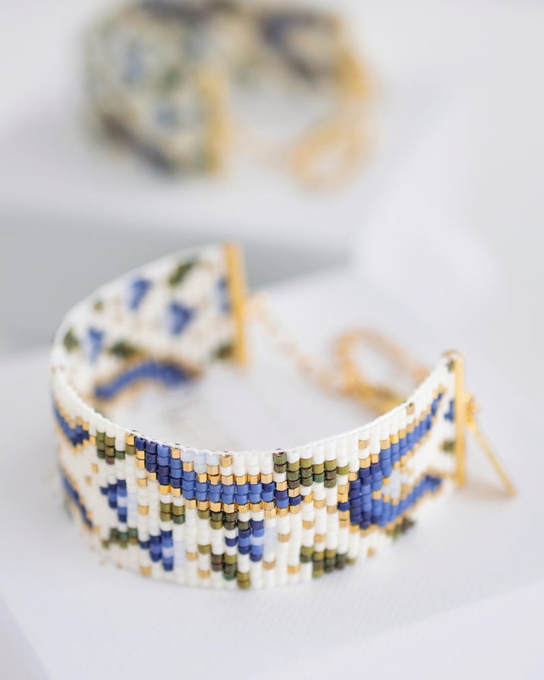May include: A beaded bracelet with a white base, featuring a geometric pattern in blue, green, and gold. The bracelet is made of small beads and has gold-colored metal clasps and chain. Another bracelet is slightly out of focus in the background.