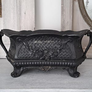 May include: A black cast iron planter with ornate floral and woven basket design. The planter has two handles and sits on four decorative feet. The planter is approximately 30 cm long and 15 cm tall.