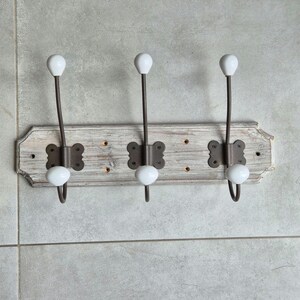 May include: A distressed wooden coat rack with three metal hooks. Each hook has a white ceramic knob and is mounted on a rectangular, off-white wooden board. The board has decorative edges.