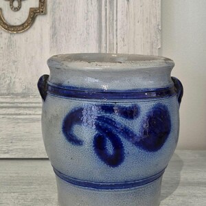 May include: A blue and grey stoneware crock with dark blue decorative accents. The antique jar has two small handles and a rounded shape. The design includes a dark blue floral pattern and horizontal bands.