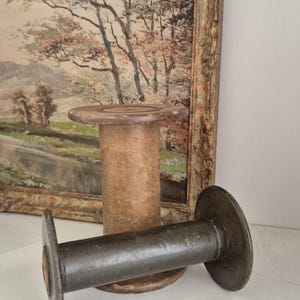 May include: Two vintage wooden spools, one standing and one lying down, with a landscape painting in the background. The spools are cylindrical with circular ends. The painting has a gold frame.