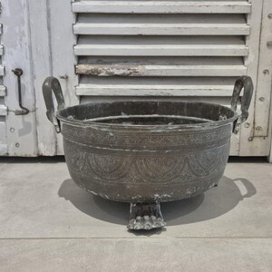 May include: A dark, weathered metal basin featuring two upright handles and three feet shaped like animal paws. The exterior surface is decorated with an intricate, repeating engraved pattern of arches and floral motifs around the entire circumference.