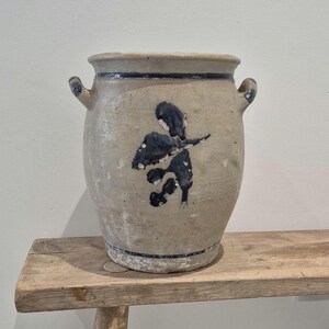 May include: A vintage stoneware crock with a light beige body and dark blue floral design. The crock has two small handles and a dark blue band around the top. It sits on a rustic wooden bench.