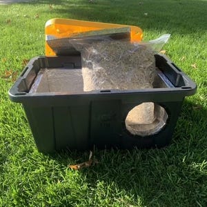 May include: A dark green plastic cat house with a round entry hole. Inside is a clear plastic bag filled with hay. A yellow lid is partially open on top. The cat house is sitting on green grass.