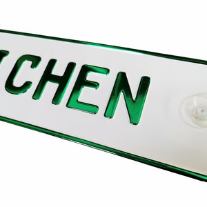 Könnte beinhalten: Eine rechteckige weiße Kennzeichentafel mit dem Wort "EICHEN" in grünen, glänzenden Buchstaben. Die Tafel hat einen grünen Rand und einen transparenten Saugnapf auf der Rückseite.