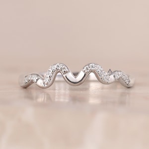 Könnte beinhalten: Ein silberfarbener Ring mit einem durchgehenden wellenförmigen oder serpentinenartigen Banddesign. Die Oberseite des Wellenmusters ist mit kleinen, klaren, funkelnden Steinen besetzt, die der geschwungenen Form des Metallbandes an der Vorderseite folgen.