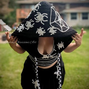 Puede incluir: Un top y una falda de crochet negro con diseños de arañas y telarañas blancas. El top tiene un escote pronunciado y la falda tiene una silueta larga y fluida. El atuendo se complementa con una capucha de crochet negro con diseños de arañas y telarañas blancas.
