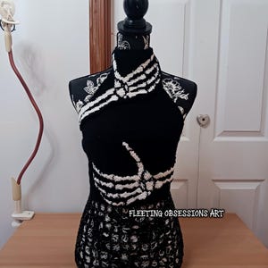 May include: A black crochet halter top featuring a white skeleton hand design across the chest and neck area. The garment is displayed on a mannequin and includes the text FLEETING OBSESSIONS ART printed in white on the lower section.