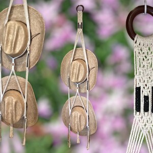 May include: A set of three macrame hat hangers with a wooden ring at the top. The hangers are made of white cord and have black accents. The hangers are hanging from a ceiling or wall and have two straw cowboy hats hanging from them.