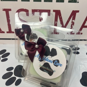 May include: Two clear wax melts in plastic containers with a white wax base. The wax melts are decorated with a burgundy bow and a pearl. The top wax melt has a white circle with black paw prints and the text "A Golden's Love Juniperberry & Pine".