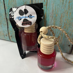 May include: Two small glass bottles filled with a red liquid, each with a wooden cap and a decorative string. One bottle is packaged in a black plastic bag with a label that reads "A Golden's Love Peppermint Milkshake". The other bottle is displayed separately.
