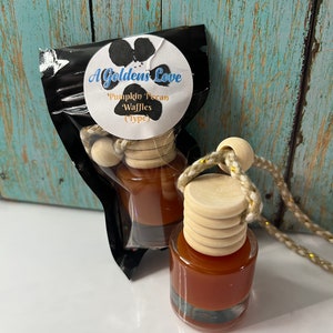 May include: A small glass bottle with a brown liquid inside, hanging from a string with wooden beads. The bottle has a wooden cap and is labeled "A Golden's Love Pumpkin Pecan Waffles (Type)".