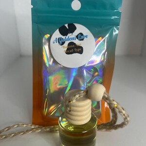 May include: A small glass bottle with a wooden cap and a gold string hanging from it. The bottle is filled with a yellow liquid. The bottle is in a clear plastic bag with a holographic design. The bag has a white circle with the text "A Golden's Love - Banana Nut Bread Type" on it.