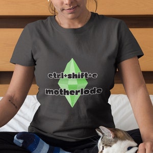 May include: Dark gray t-shirt with the text "ctrl+shift+c motherlode" and a green diamond graphic. The shirt is worn by a person sitting on a bed with a cat.
