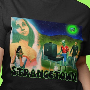 Op de afbeelding: Zwart T-shirt met een grafische print van een vrouw, een man die hotdogs grilt en een vliegende schotel. De tekst "STRANGETOWN" is in groene en zwarte letters onder de print gedrukt.