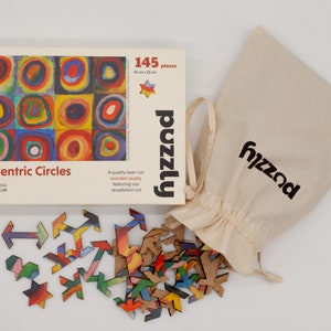 May include: A wooden puzzle with 145 pieces, featuring a colorful concentric circles design. The puzzle box is cream-coloured with the text "Concentric Circles" and "puzzly". A canvas bag with the "puzzly" logo and scattered puzzle pieces are also visible. Box size: 36 cm x 25 cm.