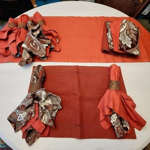 May include: A set of burnt orange table linens including a long rectangular table runner, a rectangular placemat, and several napkins. The napkins feature a brown, cream, and orange paisley pattern and are secured with woven brown napkin rings.