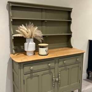 May include: A green painted wooden hutch with a top shelf and two lower cabinets. The hutch has a decorative scalloped edge and a wood top. A white vase with dried pampas grass and a glass candle jar with a wooden lid are on the top shelf.