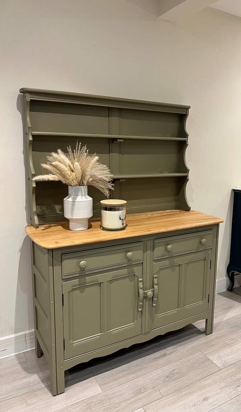 May include: A green painted wooden hutch with a top shelf and two lower cabinets. The hutch has a decorative scalloped edge and a wood top. A white vase with dried pampas grass and a glass candle jar with a wooden lid are on the top shelf.